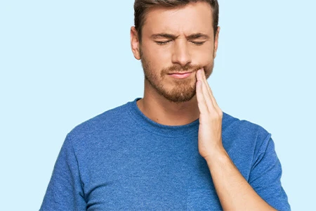 Man in a blue shirt holding his cheek, showing discomfort from tooth pain against a light blue background.